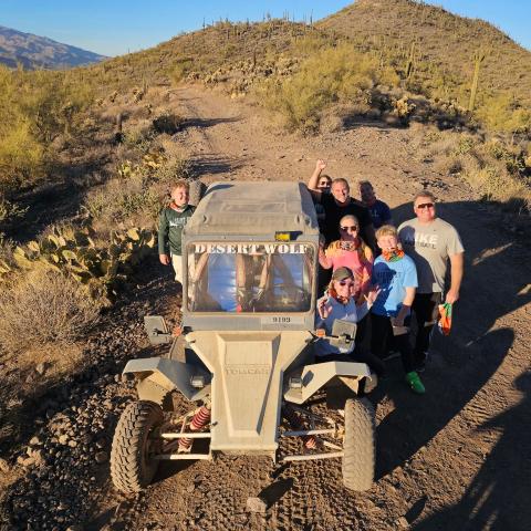 Group posing with an off-road vehicle on a desert trail surrounded by hills and cacti.