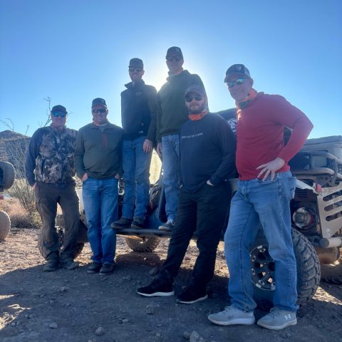 Six men posing in front of an off-road vehicle on a sunny day.