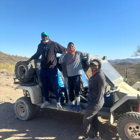 Three people standing by an off-road vehicle in a desert landscape under a clear blue sky.