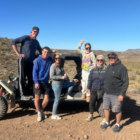 Group of six people pose in front of an off-road vehicle in a desert landscape.