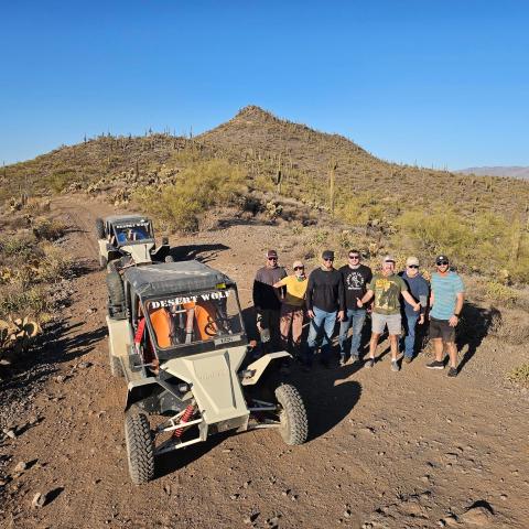 Group of people posing by off-road vehicles on a desert trail with hills in the background.