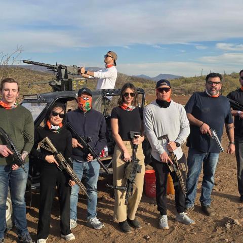 Eight people with rifles pose in front of an off-road vehicle in a desert landscape.