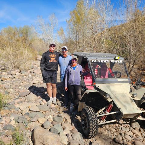 Three people stand beside a 'Desert Wolf' off-road vehicle on a rocky terrain with trees.