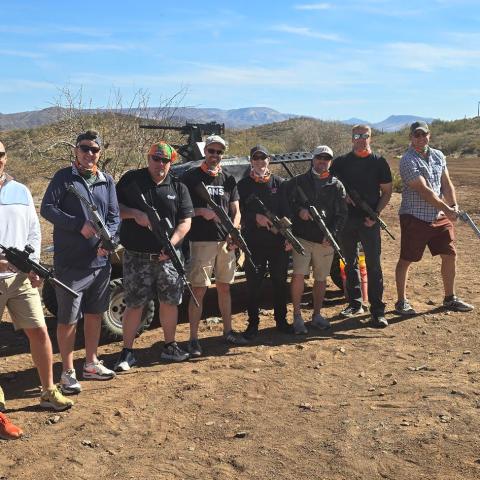 Group of people holding rifles in a desert setting with mountains in the background.