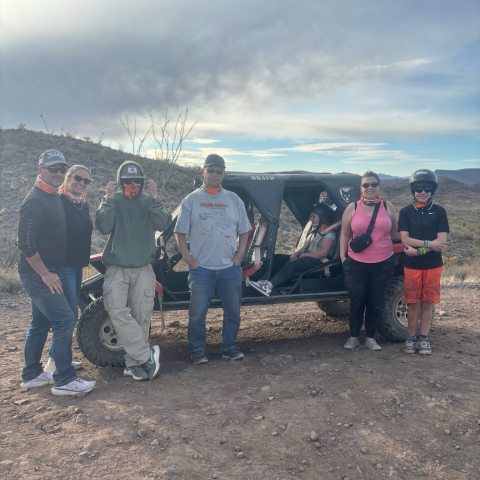 Group of six people with a dune buggy on a desert trail under a cloudy sky.