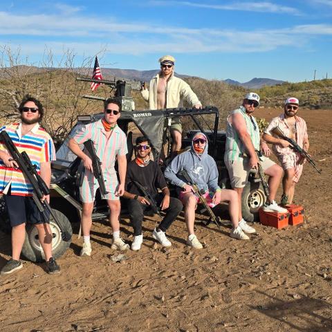 Group of seven people with firearms posing near a vehicle in a desert landscape.