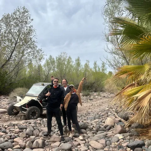 Three people pose on rocky terrain with an off-road vehicle, trees and cloudy sky in background.