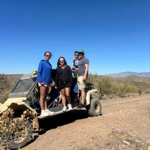 Four people stand on a parked off-road vehicle in a desert landscape under a clear blue sky.