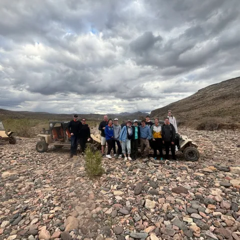 Group of people standing on a rocky terrain with off-road vehicles under a cloudy sky.