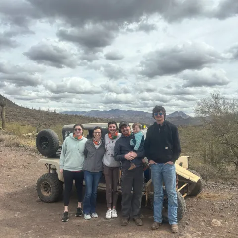 Group of six people with a baby standing in front of an off-road vehicle in a desert landscape.