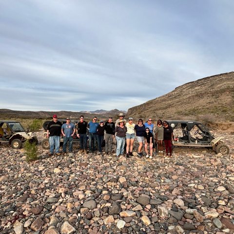 Group of people standing on rocky terrain with off-road vehicles and hills in the background.