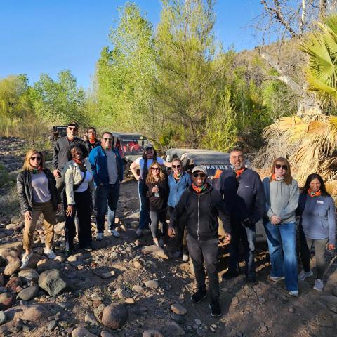 Group of people posing on a rocky trail with greenery and a vehicle in the background.