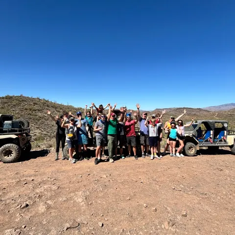 Group of people posing on a rocky terrain with jeeps under a clear blue sky.