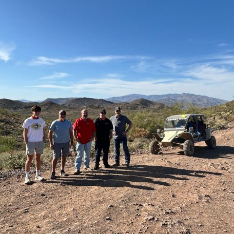 Five people standing next to a dune buggy on a desert trail with mountains in the background.