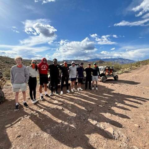 Group of people standing on a rocky desert trail with two off-road vehicles under a cloudy sky.