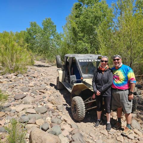 Two people stand by an off-road vehicle on a rocky path with trees in the background.