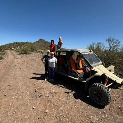 Four people and a desert buggy on a gravel road with a clear blue sky.