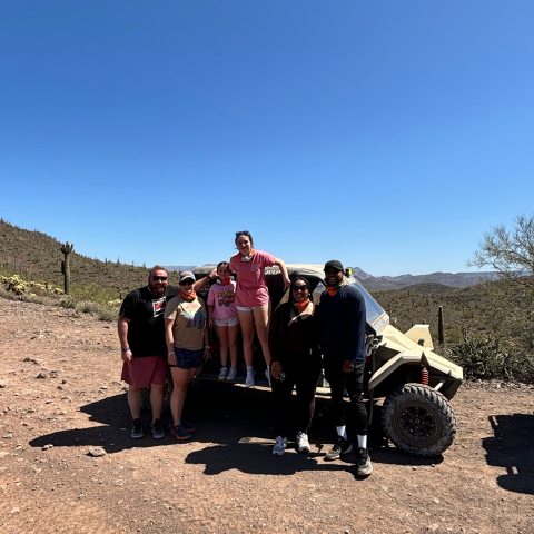 Group of people standing near an off-road vehicle in a desert landscape under a clear blue sky.