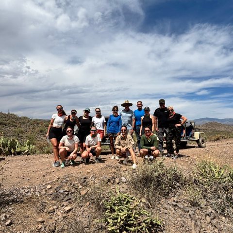 Group of people posing on a desert trail with a jeep, under a partly cloudy sky.