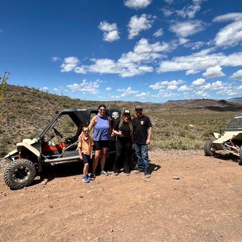 Four people standing between two off-road vehicles in a desert landscape with blue sky and clouds.