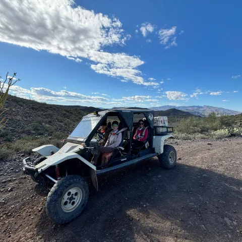 Off-road buggy with two people in desert landscape under a blue sky with clouds.