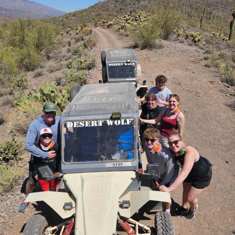 Group of people with dune buggy on a desert trail, surrounded by cacti and hills under a clear blue sky.