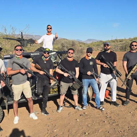 Nine people pose with rifles in a desert with off-road vehicles and mountains in the background.