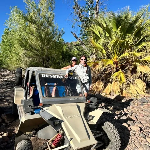 Two people posing with a dune buggy in a rocky desert landscape with trees.