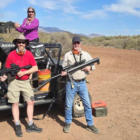 Three people with guns pose by an off-road vehicle in a desert landscape under a blue sky.