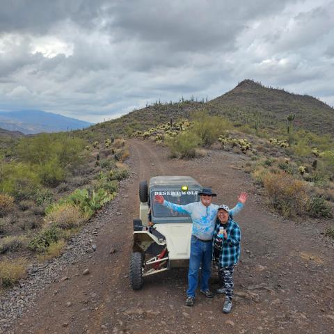 Two people in front of an off-road vehicle on a desert trail with cacti and cloudy sky.