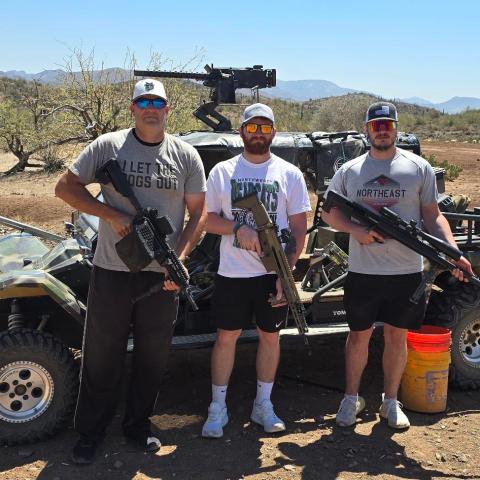 Three people with guns stand in front of an ATV in a desert landscape.