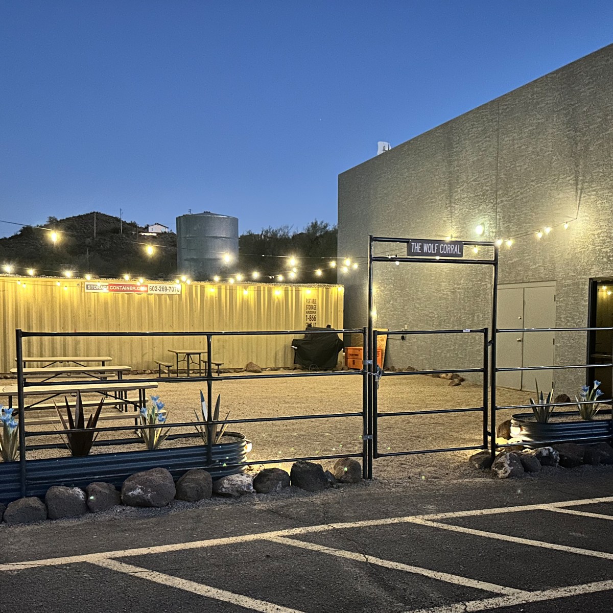 Outdoor area with string lights, benches, and a sign reading 'The Wolf Corral' at dusk.
