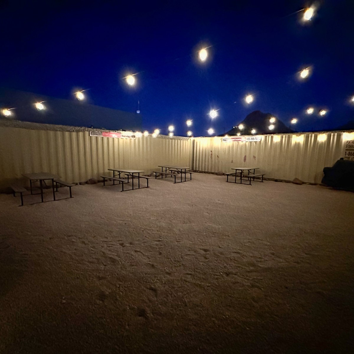Outdoor area with string lights, empty benches, and shipping containers at night.